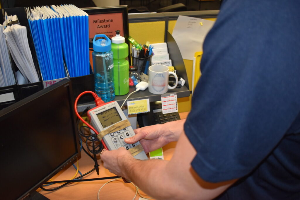 Technician performing Portable Appliance Testing (PAT) using a testing device on a desk with office supplies, emphasizing workplace safety and compliance with Australian standards.