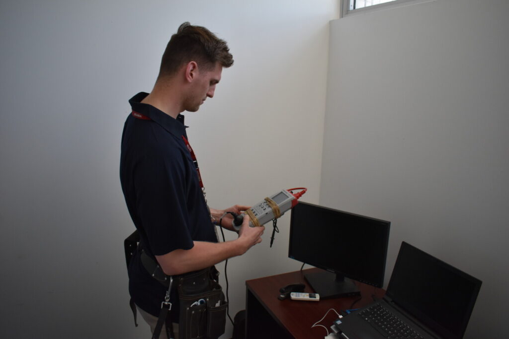 Technician conducting electrical appliance testing using a testing device, with a computer monitor and laptop on a desk, emphasizing workplace safety and compliance with AS/NZS 3760 standards.